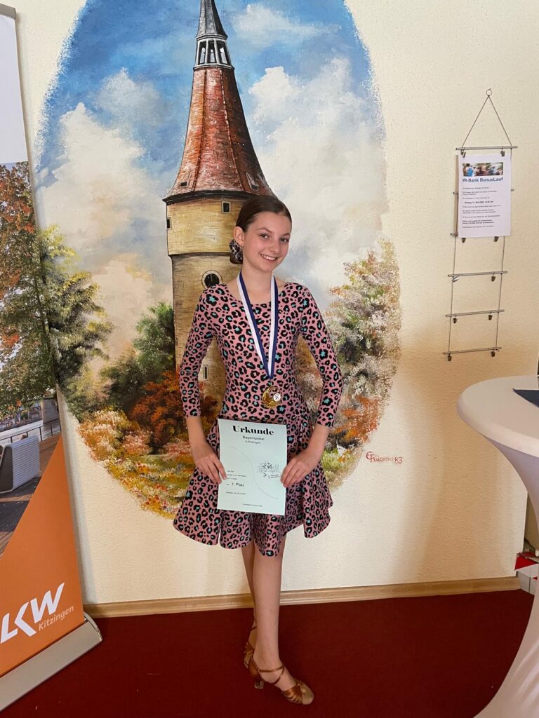Sofie steht mit der Urkunde in der Hand vor einer Wand mit einem Gemälde im Hintergrund