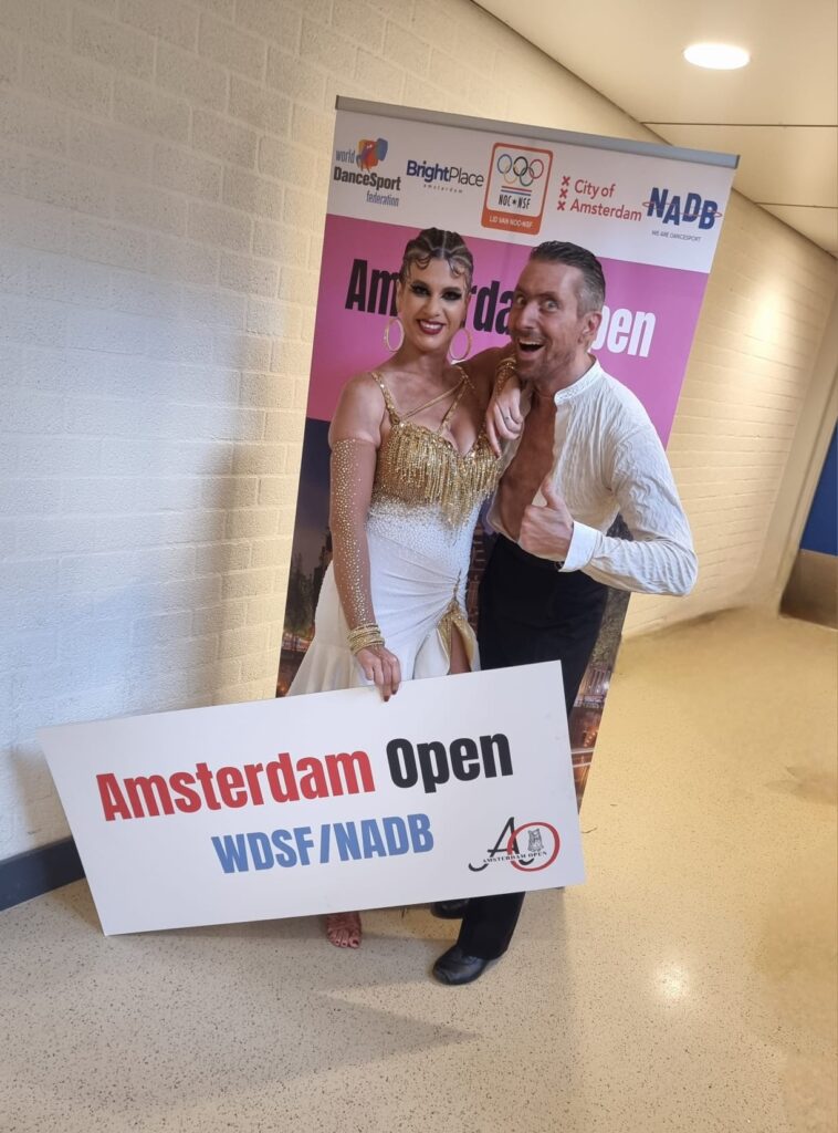 Kirsten und Bernhard stehen vor dem Plakat mit "Amsterdam Open" und lachen in die Kamera